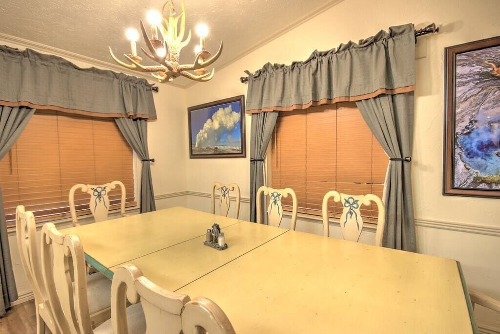A dining room with a long table, six chairs, a chandelier, and two windows with curtains.