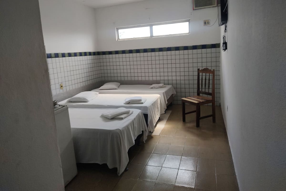 A small, clean room with three beds, a wooden chair, and a window providing natural light.