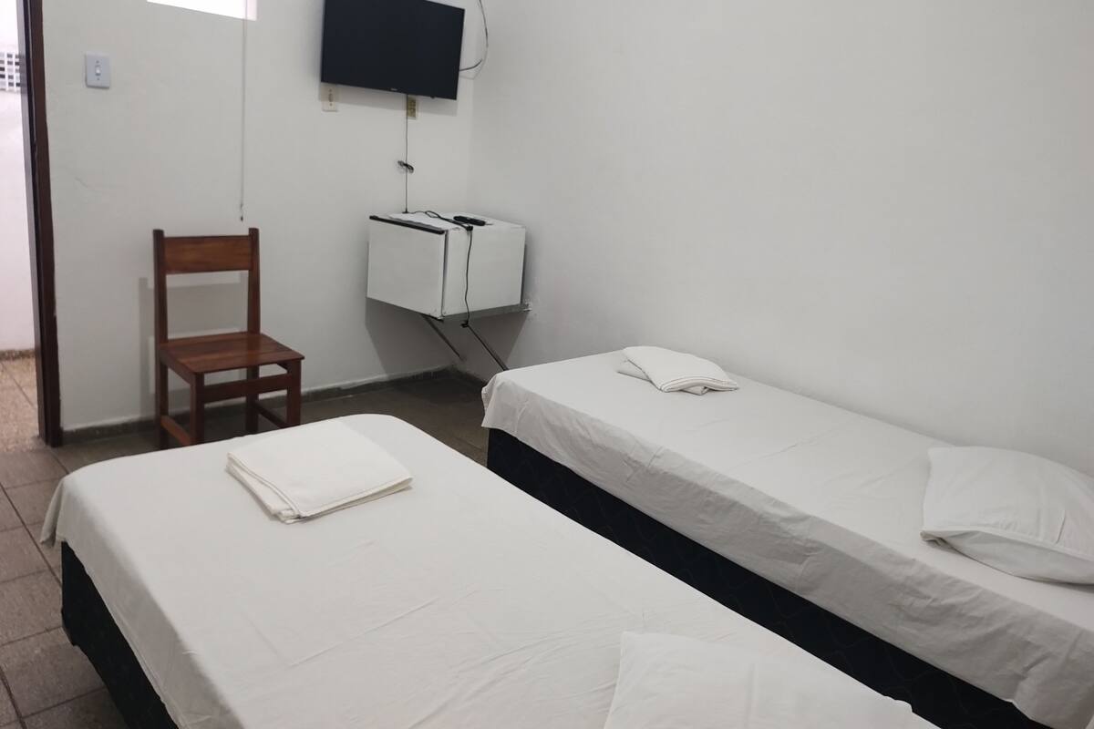 A simple room with two beds, a chair, a TV mounted on the wall, and a small white cabinet.