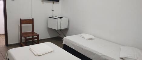 A simple room with two beds, a chair, a TV mounted on the wall, and a small white cabinet.