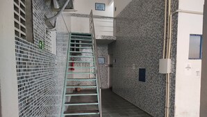 Un corridoio stretto con una scala in metallo che conduce a un livello superiore. Le pareti sono rivestite di mosaici e ci sono cartelli che indicano indicazioni e numeri di stanza.
