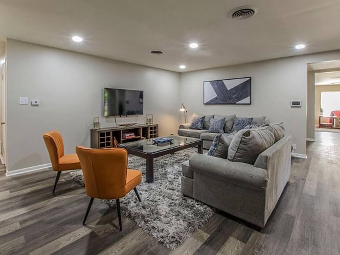 Een moderne woonkamer met een grijze bank, oranje fauteuils, een salontafel en een flatscreen televisie aan de muur.