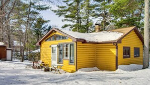 A yellow cabin with a gabled roof, surrounded by snow and trees. - Boat Dock & Fire Pit: Waterfront Home in Crosslake (Crosslake)
