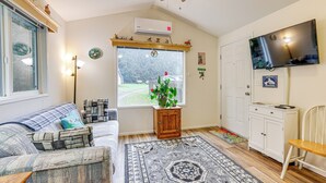 A cozy living room with a sofa, a small wooden cabinet, a television, and a window with a view of a garden. - Remote Workers Welcome: Tiny Home in Port Angeles! (Port Angeles)