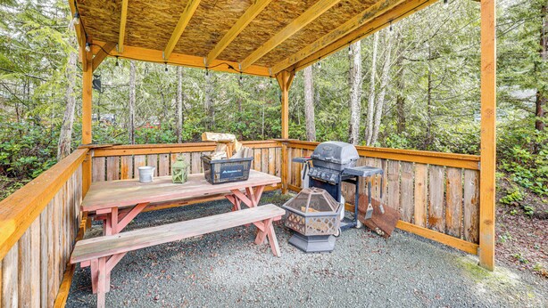 A covered wooden deck with picnic tables and a grill, surrounded by forest. - Remote Workers Welcome: Tiny Home in Port Angeles! (Port Angeles)