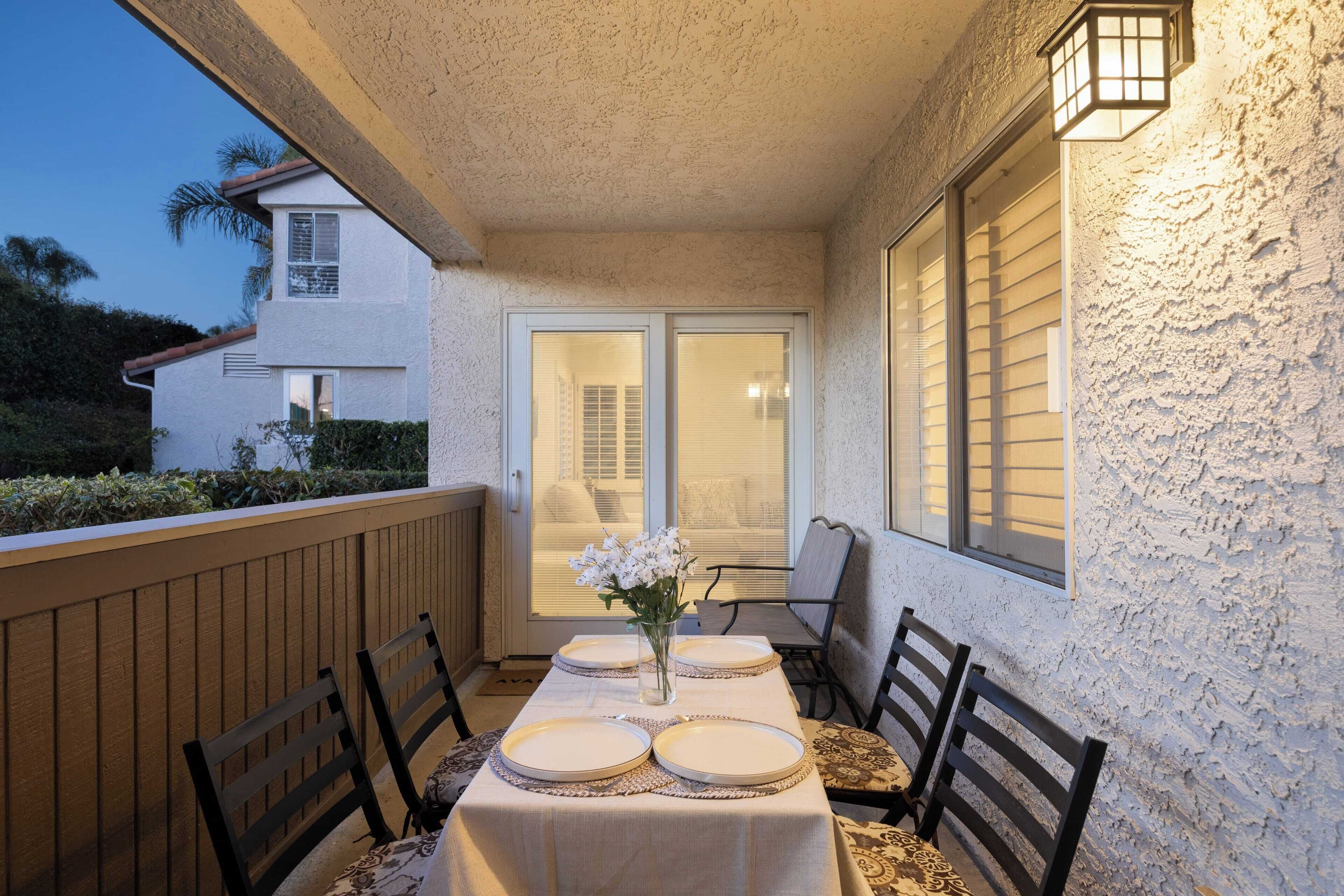 A dining area with a table set for four, chairs, a vase with flowers, and a view of a house through a glass door.
