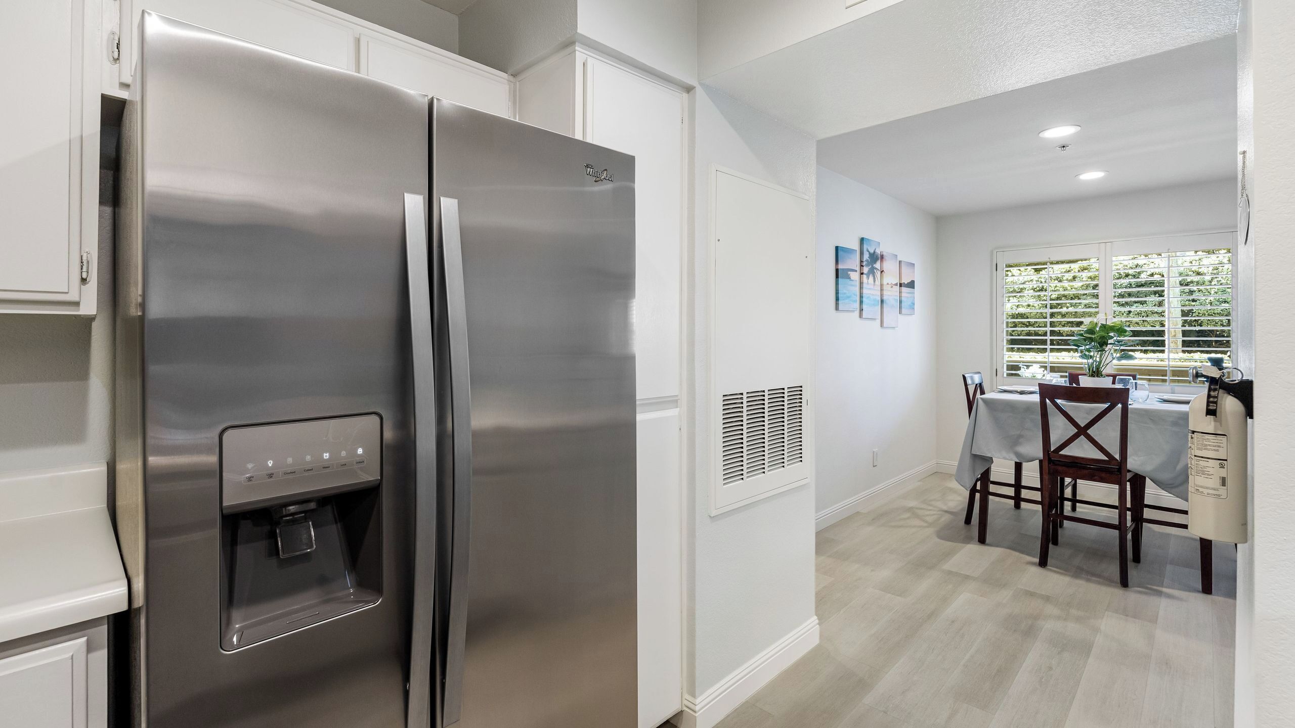 A stainless steel refrigerator with a water dispenser, a dining area with a table and chairs, and a window with blinds.