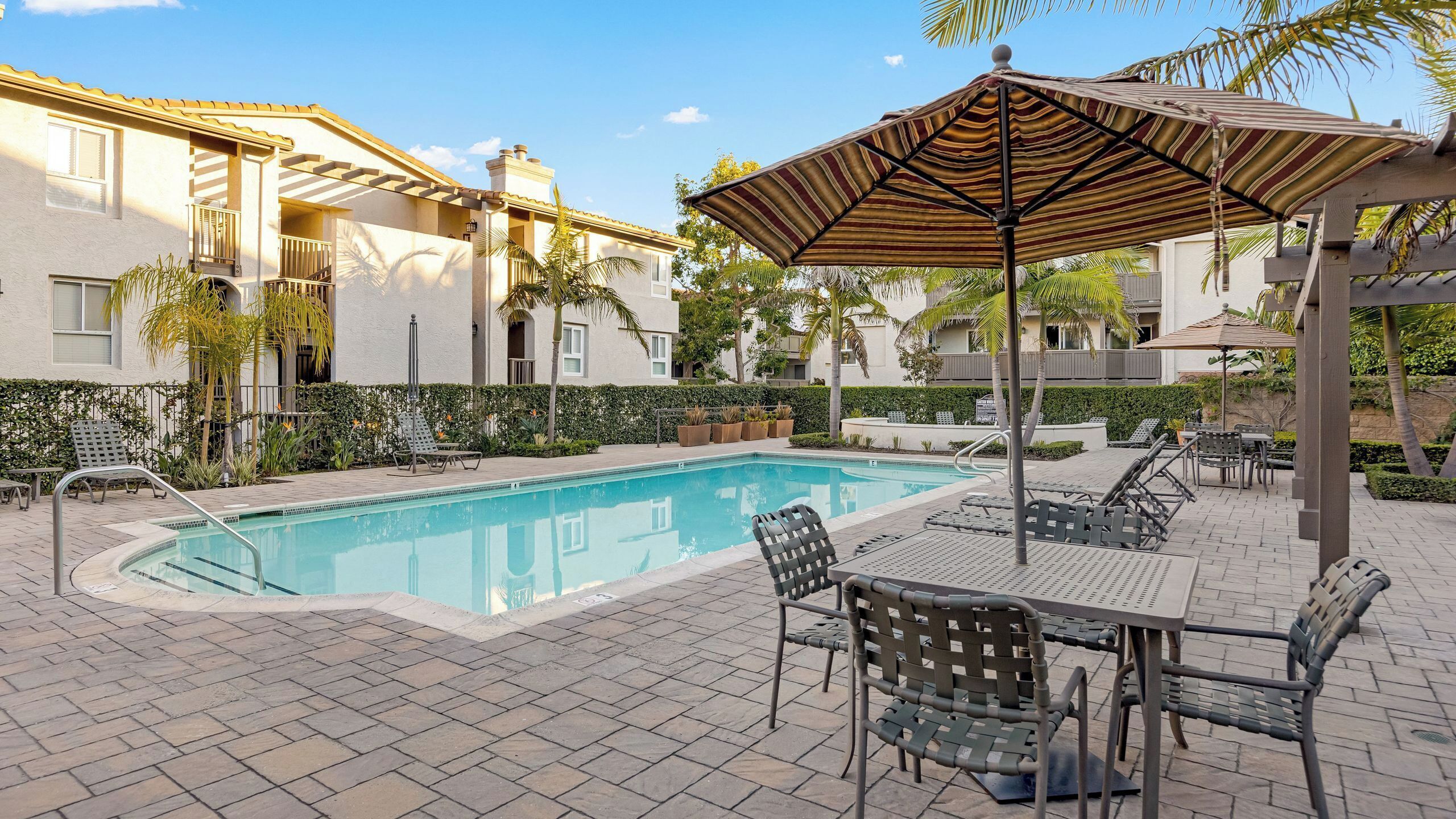 A swimming pool with lounge chairs and umbrellas, surrounded by a paved patio and greenery.