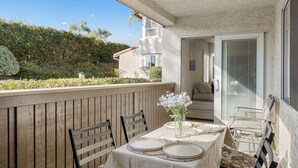 Un balcone con tavolo apparecchiato per consumare pasti, sedie, un vaso con fiori e vista su una casa e vegetazione.
