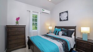 Une chambre à coucher comprenant un lit, des tables de chevet, une commode, une fenêtre avec des rideaux, un appareil de climatisation et une œuvre d’art accrochée au mur.