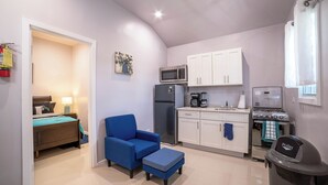 Un espace de vie compact comprenant un coin cuisine, un lit avec une chaise et un pouf bleus, et une poubelle.