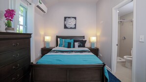 Une chambre à coucher comprenant un lit, des tables de chevet, une commode, un climatiseur, une fenêtre avec des rideaux, un tableau représentant des fleurs accroché au mur et une porte de salle de bain.