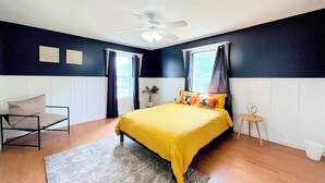 Une chambre à coucher avec un lit, une chaise, un ventilateur de plafond et une fenêtre avec des rideaux.
