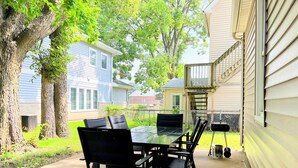Une terrasse extérieure avec une table en verre et des chaises noires, entourée d’arbres et d’une clôture.