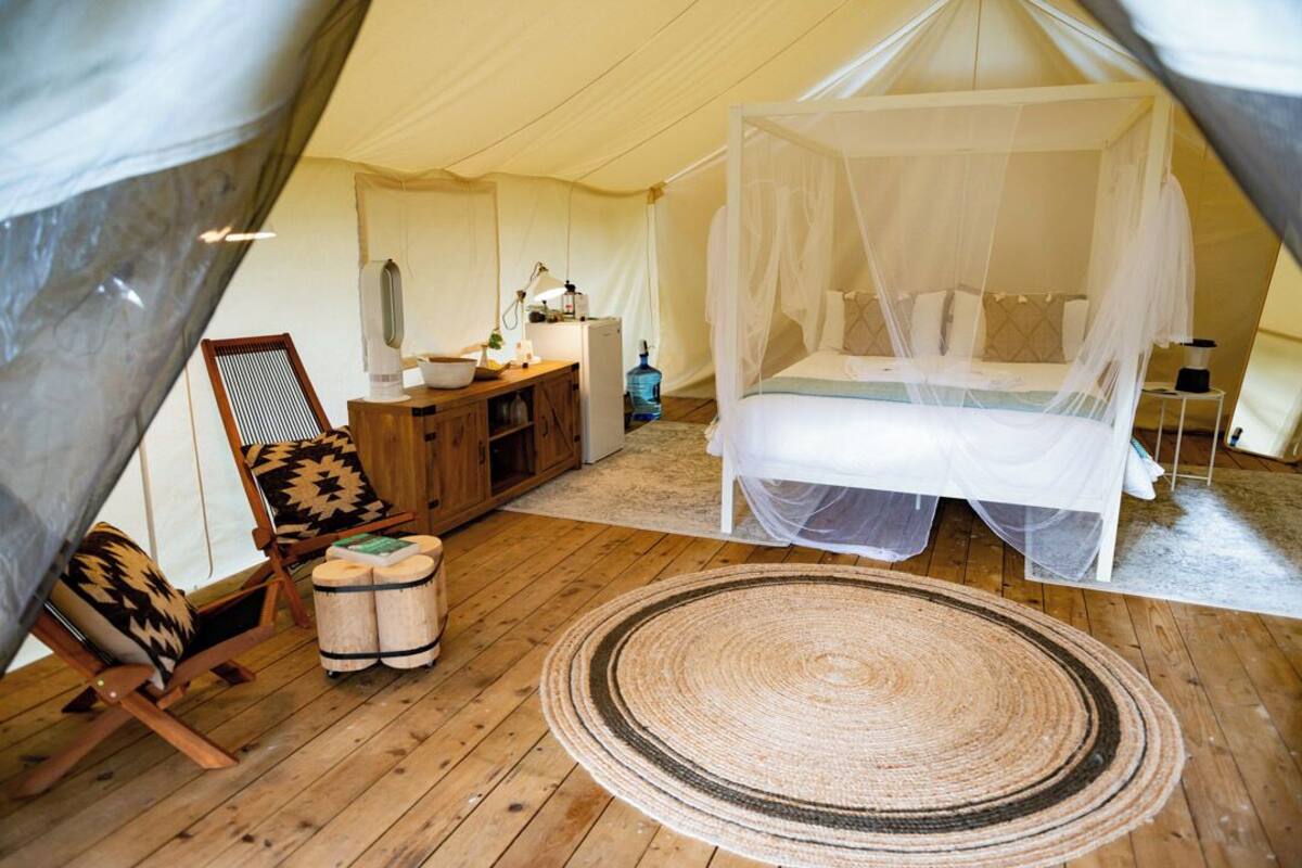 A tented accommodation with a bed under a canopy, a wooden chair, a small table, and a rug.