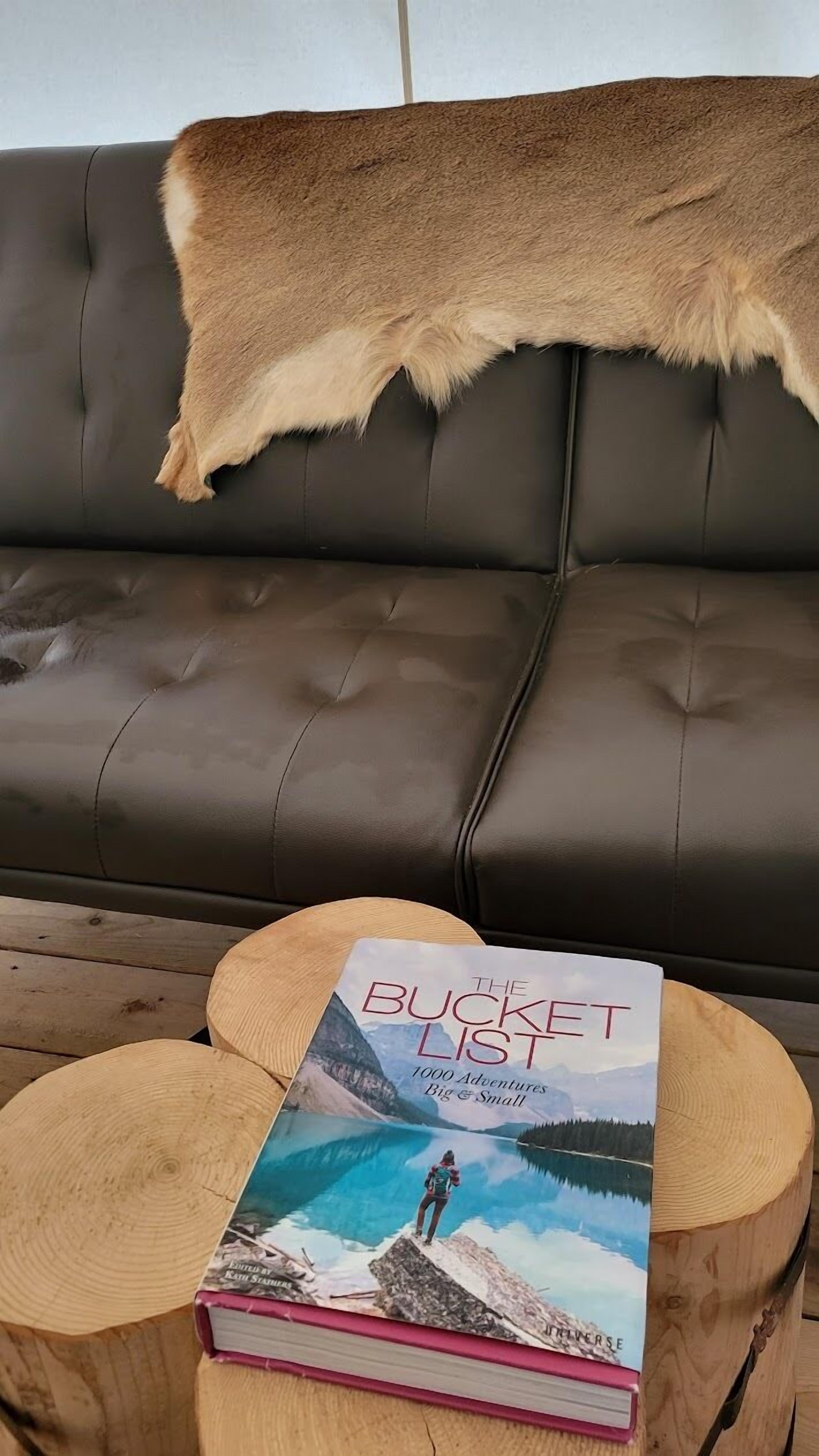 Un sofá de cuero con una manta de piel, un libro titulado 'The Bucket List' sobre un taburete de madera y una mesa de madera.