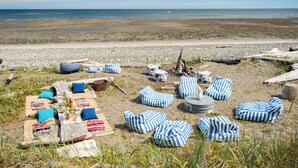 Un montaje en la playa con cojines a rayas y una mesa, rodeado de rocas y madera a la deriva.