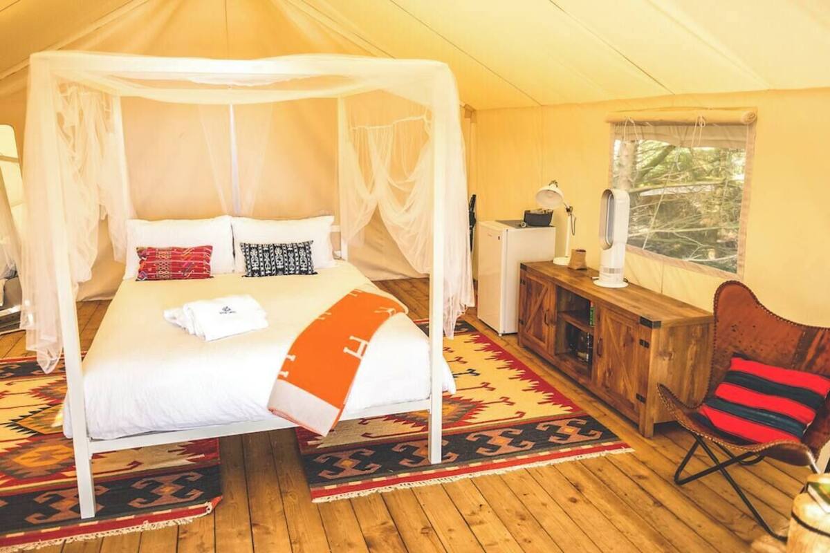 A bedroom with a four-poster bed, a wooden cabinet, a chair, and a window with a view of greenery.
