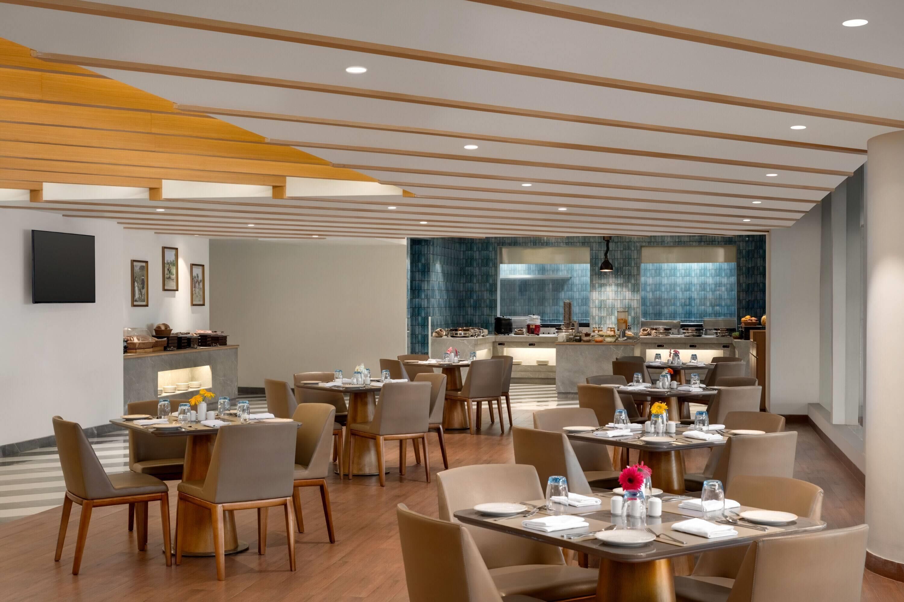 A modern dining area with multiple tables and chairs, a kitchen in the background, and a ceiling with wooden beams.