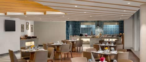 A modern dining area with multiple tables and chairs, a kitchen in the background, and a ceiling with wooden beams.