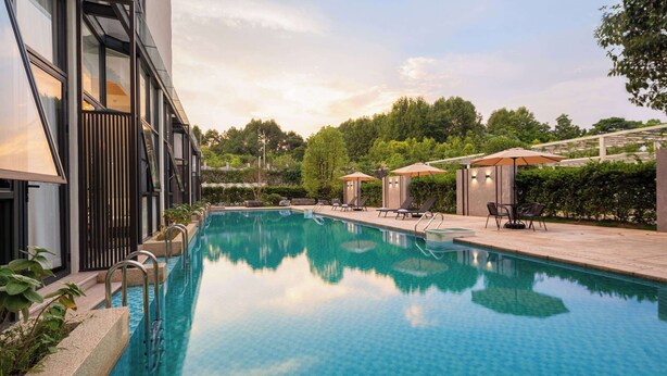 Una piscina exterior moderna con agua azul cristalina, rodeada de sillones de descanso y sombrillas, con árboles y un edificio con amplias ventanas al fondo.