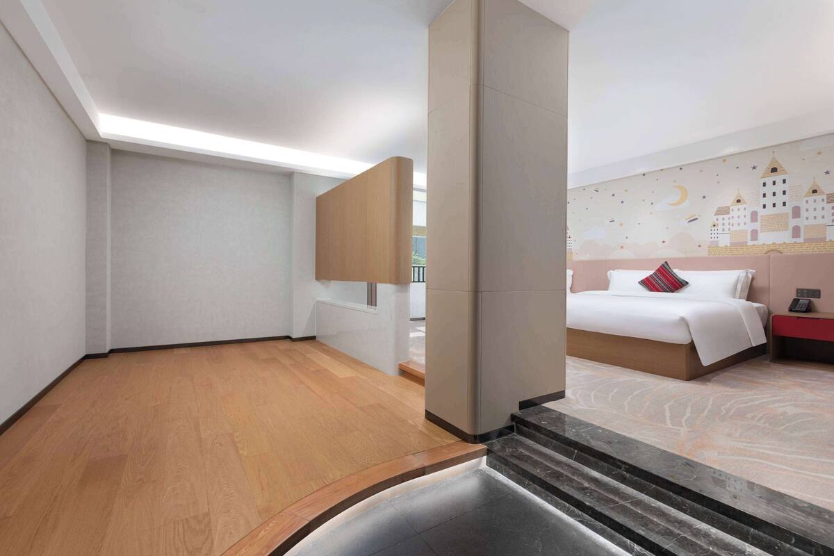 A hotel room with a wooden floor, a bed with white bedding and a red pillow, a nightstand with a phone, and a wall with a cityscape mural.