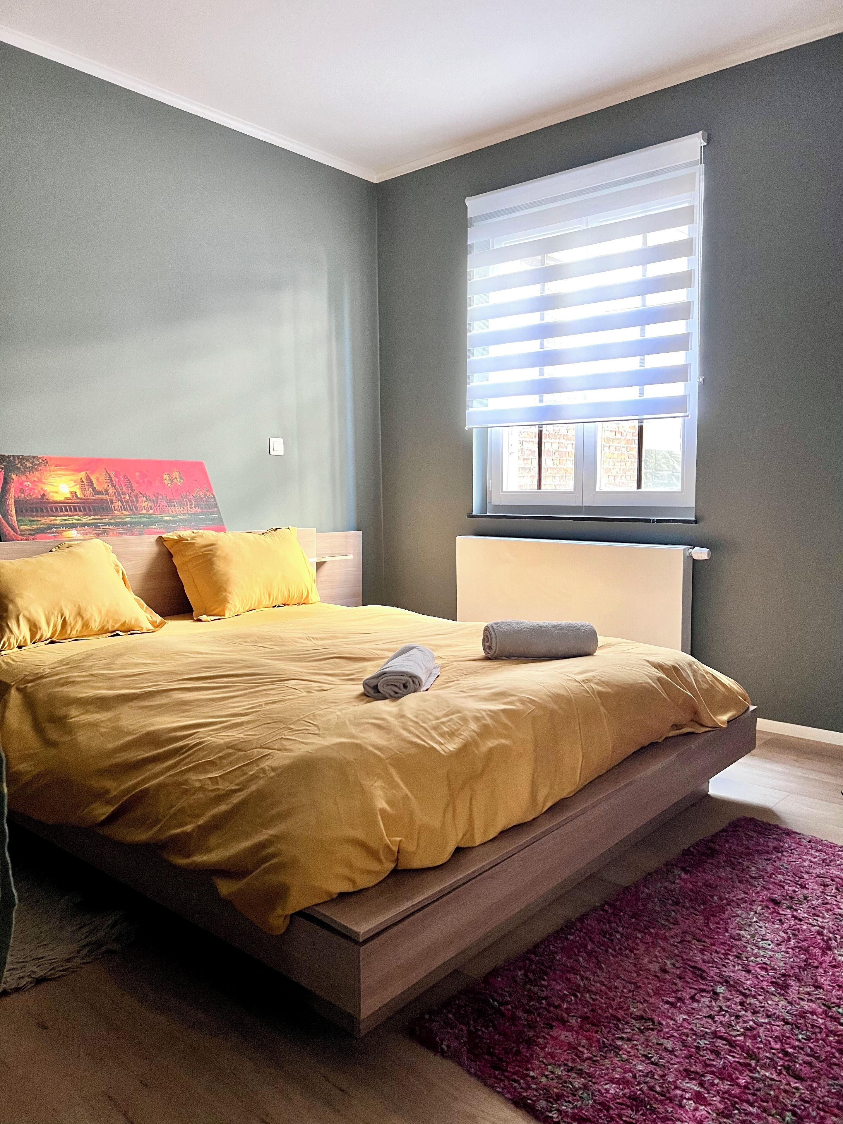 A modern bedroom with a bed, a window with blinds, a painting on the wall, and a purple rug.