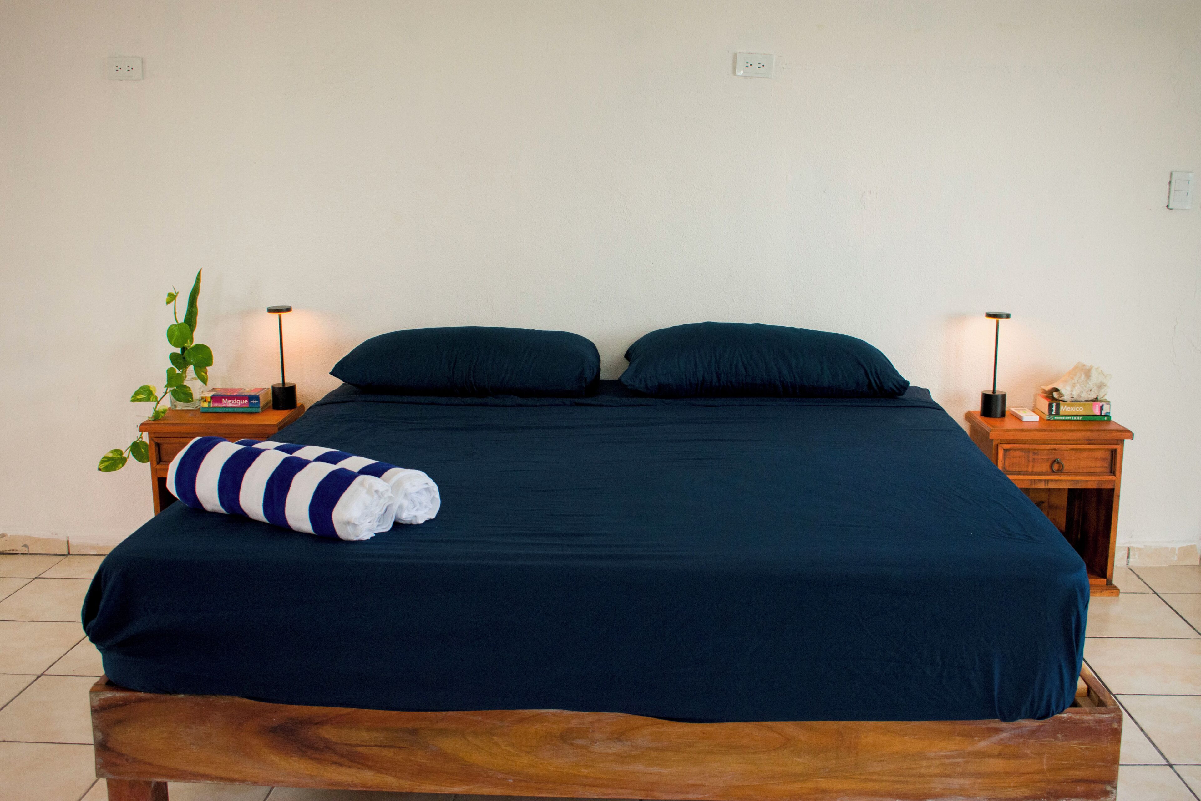Una cama con colcha y almohadas azul oscuro, una mesita de noche de madera con una lámpara, una planta y una toalla doblada.