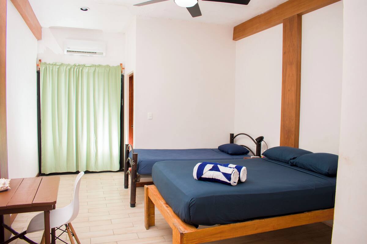 A room with two beds, a wooden table, a white chair, and a ceiling fan.