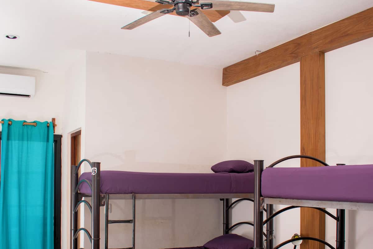 A dormitory room with bunk beds, a ceiling fan, wooden beams, and a teal curtain.