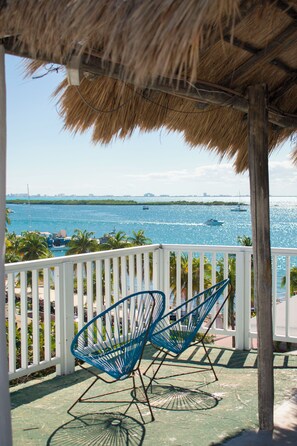 A balcony with a thatched roof, two blue wire chairs, and a view of the sea with boats. - Balu Hotel   (Isla Mujeres)