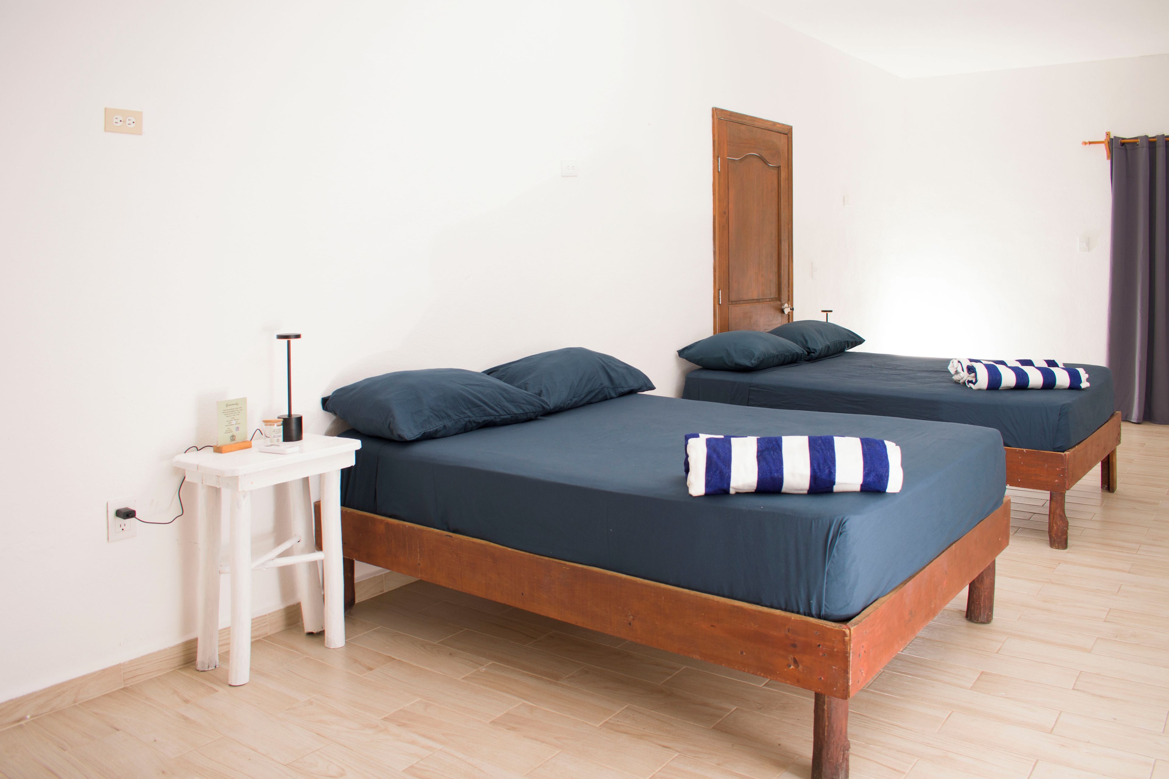 Quarto com duas camas de madeira, cada uma com colchão e almofadas azul-escuro. Há uma mesa de cabeceira branca com uma luminária e um pequeno objeto. O quarto possui piso e porta de madeira.