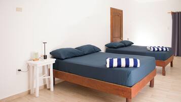 Quarto com duas camas de madeira, cada uma com colchão e almofadas azul-escuro. Há uma mesa de cabeceira branca com uma luminária e um pequeno objeto. O quarto possui piso e porta de madeira.