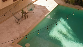 A swimming pool with a wooden deck, two chairs, and string lights.