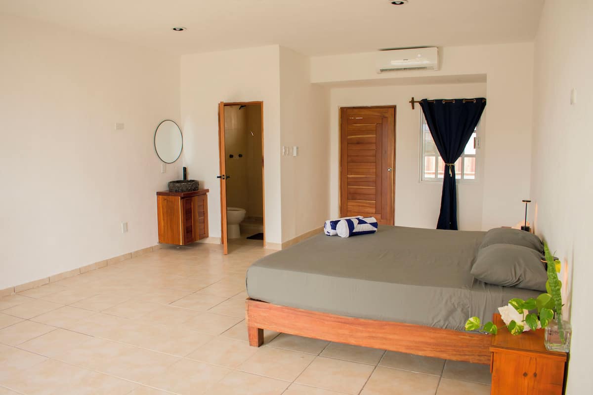 A bedroom with a bed, a wooden nightstand, a mirror, a door leading to a bathroom, and a window with a curtain.