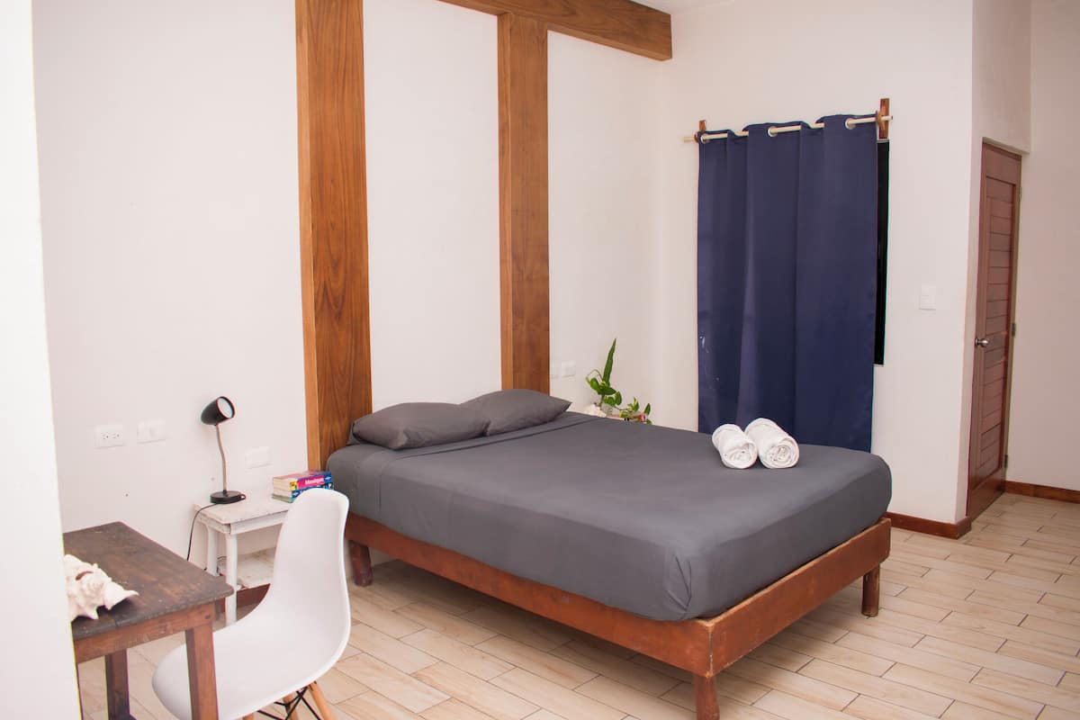 A bedroom with a wooden bed frame, a desk with a chair, a small table, and a blue curtain.