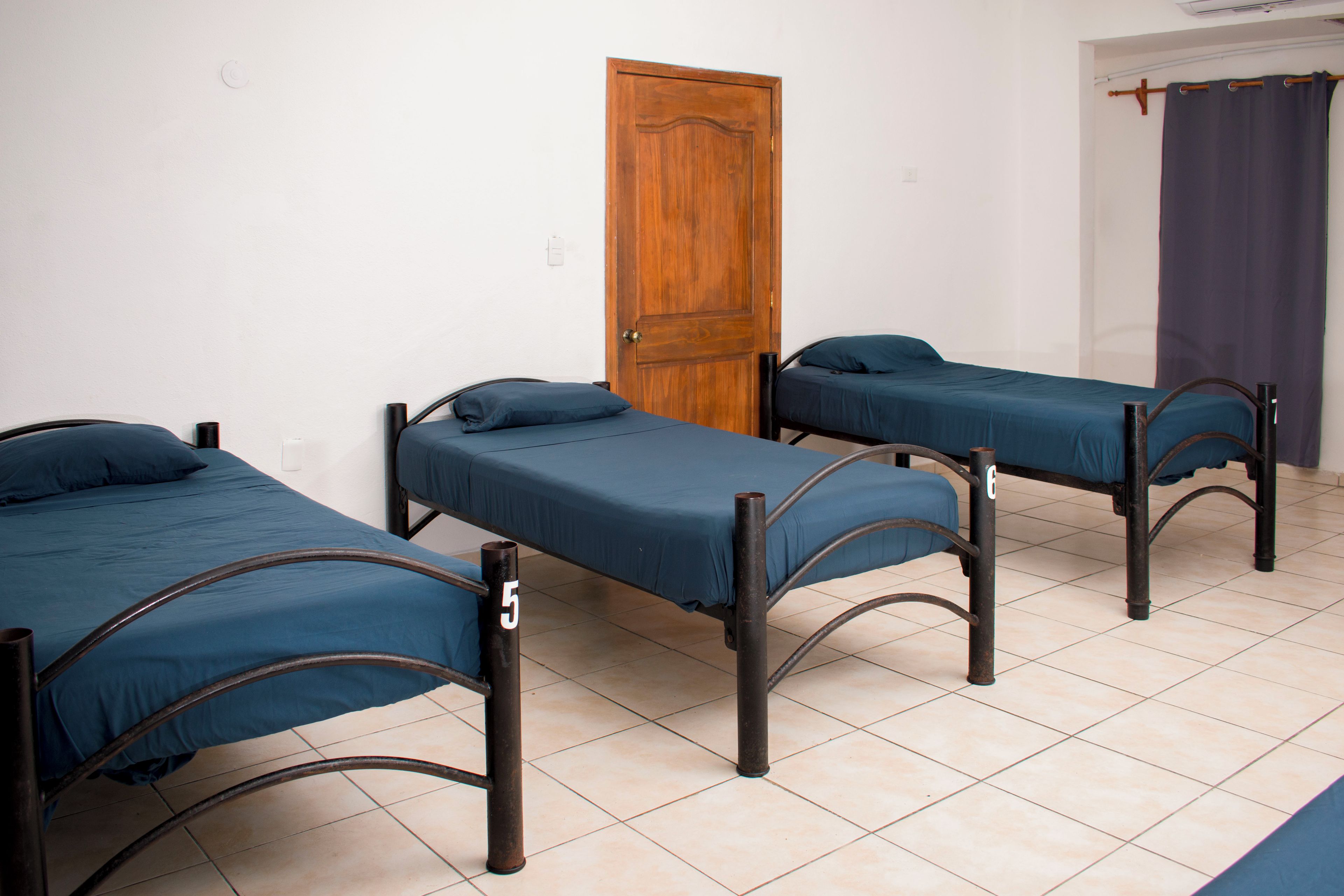 A room with three single beds, each with a number tag, a wooden door, and a curtain on the right.