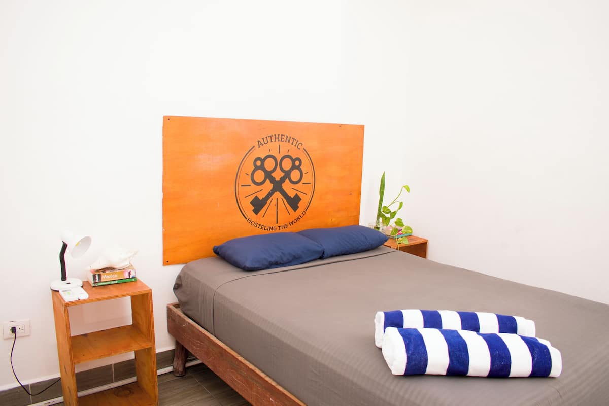 A neatly made bed with a blue pillow, a striped towel, and a wooden nightstand with a lamp and books.