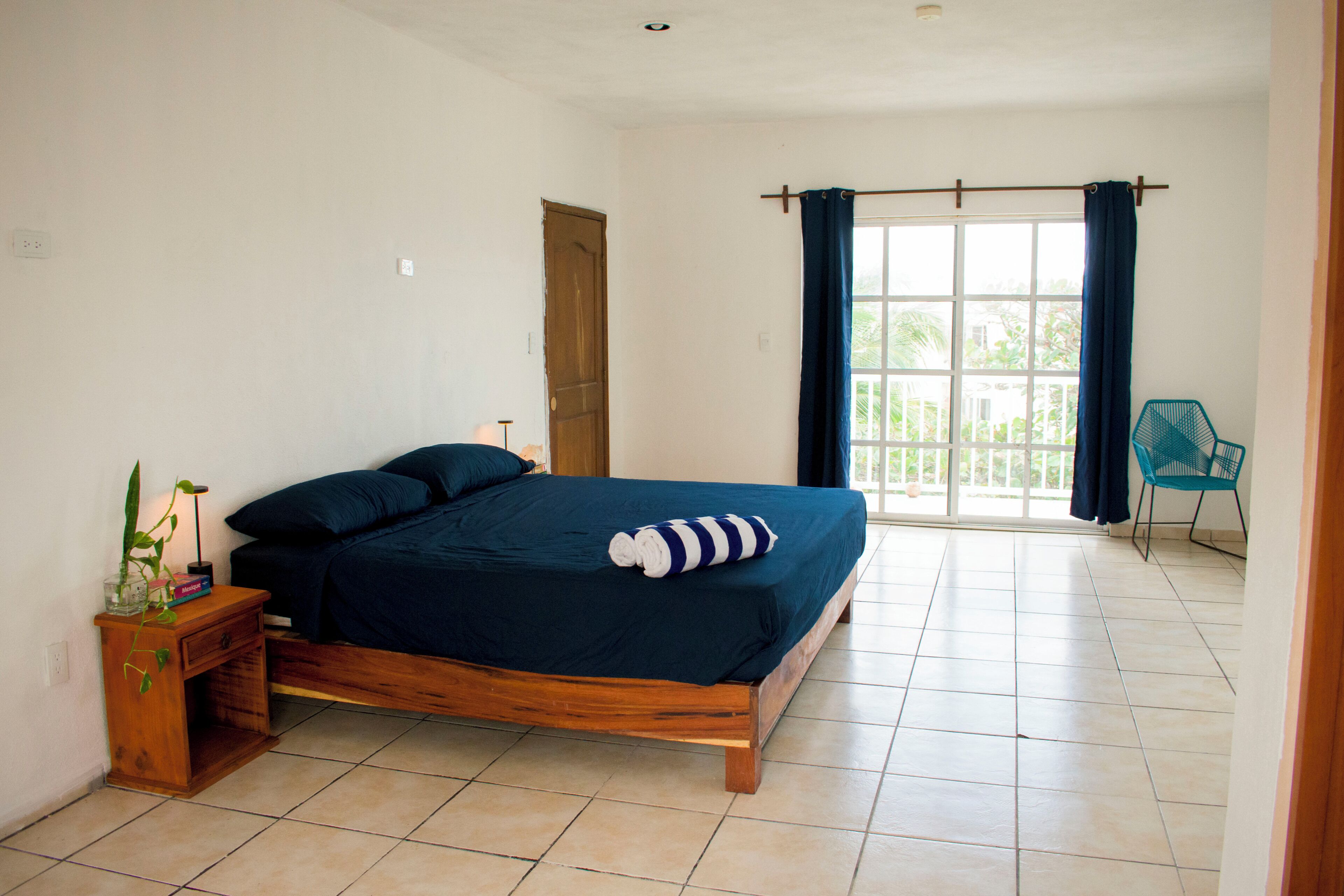 A bedroom with a wooden bed, a nightstand with a plant, a window with curtains, and a chair.