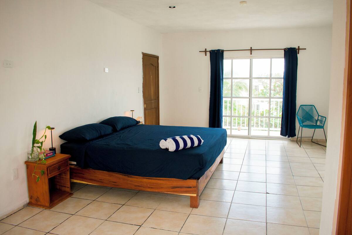 A bedroom with a wooden bed, a nightstand with a plant, a window with curtains, and a chair.