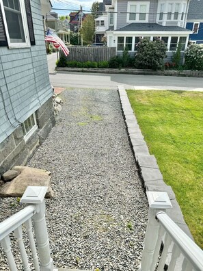 A residential driveway with a gravel path, a wooden fence, and a grassy lawn. - Captain`s Quarters in the Heart of Bar Harbor (Bar Harbor)