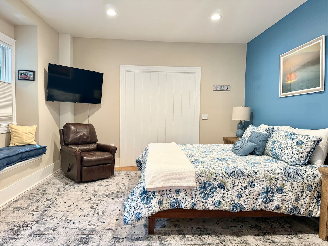 A bedroom with a bed, a recliner, a TV, and a window with blinds.