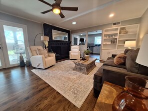A spacious living room with a large window, a ceiling fan, and a fireplace. - Heated Pool, Golf & Poker Room (Dallas)