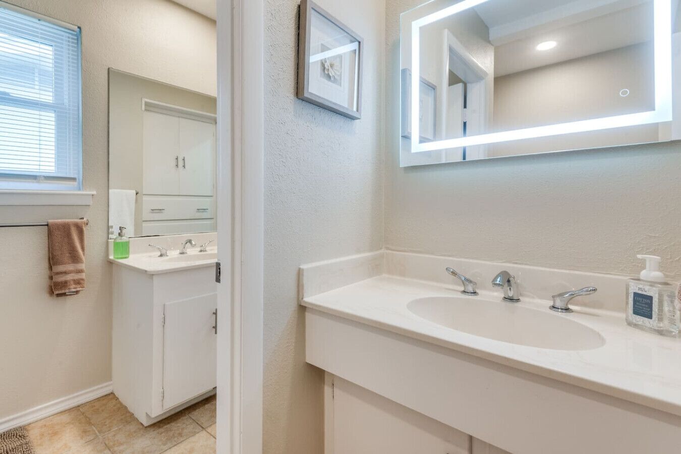 Une salle de bain avec un lavabo blanc, deux robinets, un miroir avec un éclairage intégré et un porte-serviettes.