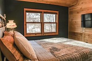 Une chambre avec un plafond en bois, une grande fenêtre donnant sur les arbres, un lit avec de nombreux oreillers et un téléviseur fixé au mur.