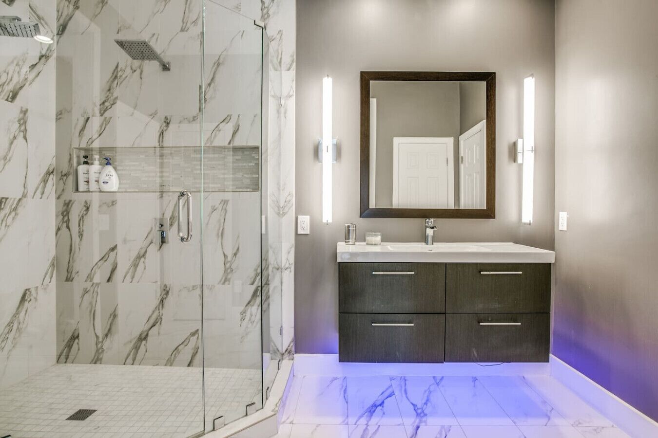 Un baño moderno con ducha envuelta en vidrio, suelo de mármol, tocador de madera oscura y un espejo grande con luces empotradas.