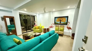 A modern living room with a teal sofa, a green armchair, and a dining area with a table and chairs. There is a large window with a view of the outdoors. - Charming, fully appointed 1-bedroom apartment in PV’s amazing Zona Romantica (Puerto Vallarta)