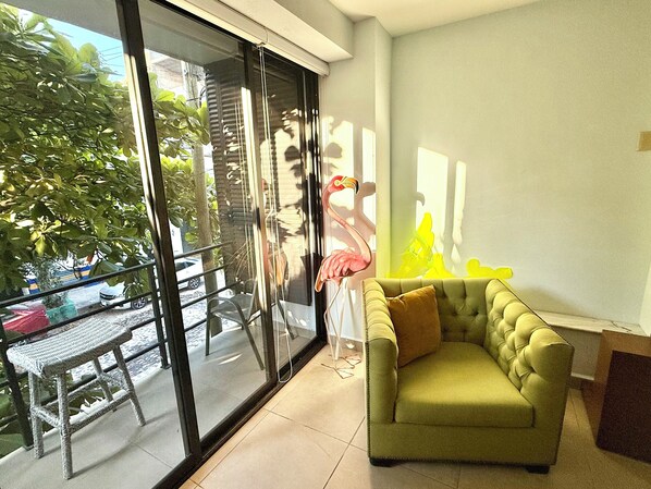 Un salon avec un canapé vert, une porte vitrée donnant sur l’extérieur et une vue sur une rue bordée d’arbres et où circulent des voitures.