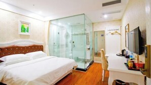 Habitación de hotel con cama, escritorio, televisión y zona de ducha con paredes de vidrio.