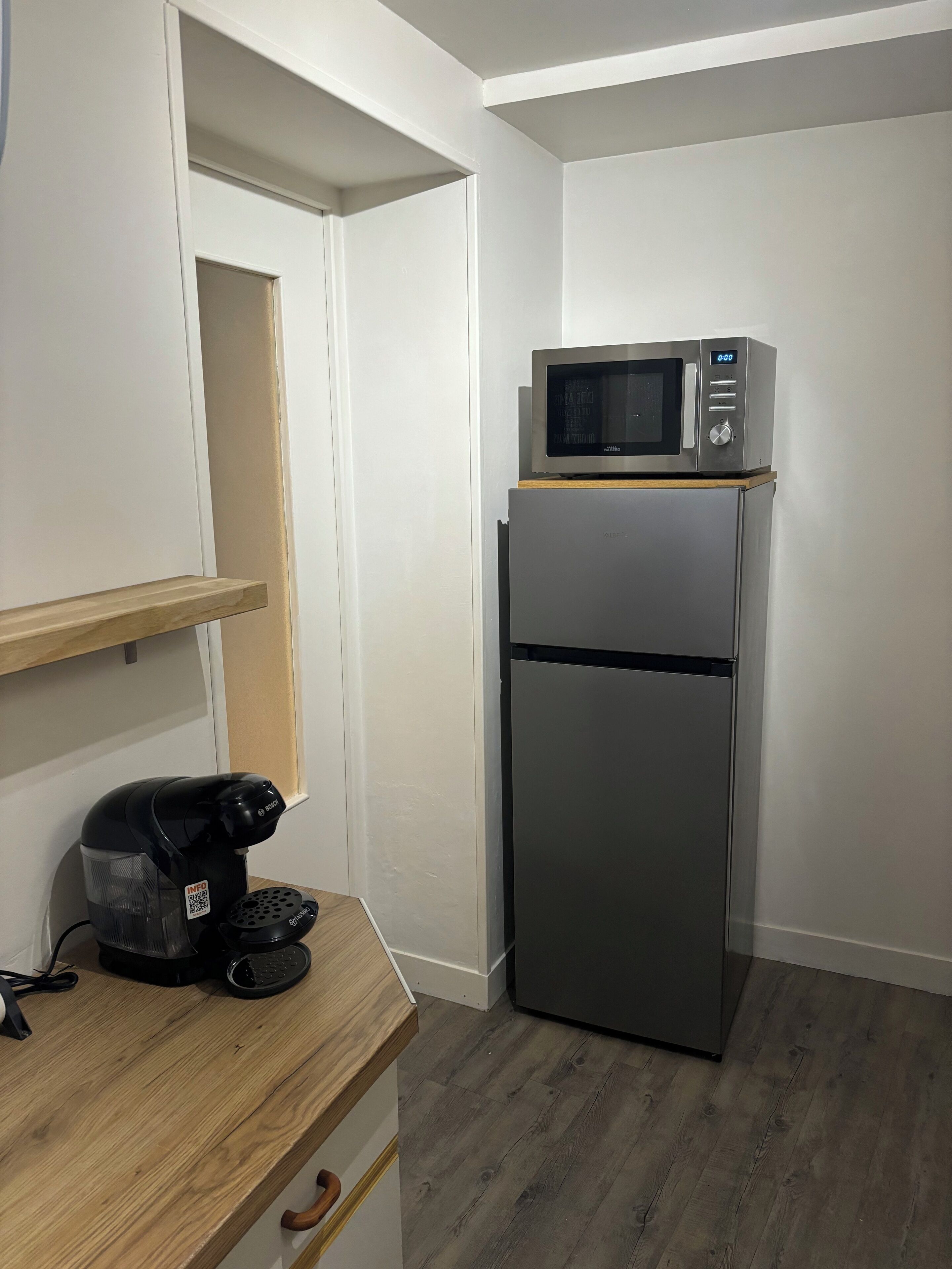 Une petite cuisine équipée, avec un micro-ondes posé sur le dessus d’un réfrigérateur en acier inoxydable, une machine à café sur un plan de travail en bois et une porte entrouverte.